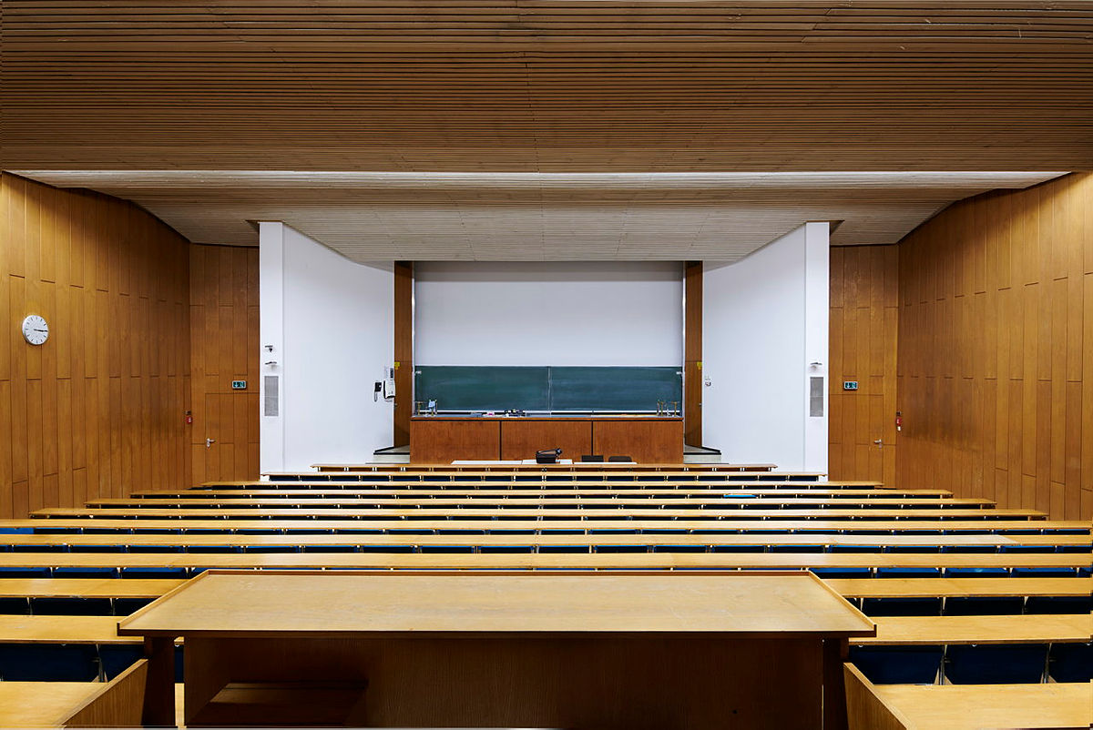 Leerstehender Hörsaal mit Reihen aus Holzbänken und Tafel an der Wand.
