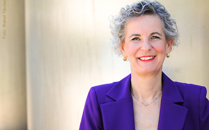 Person mit grauem kurzem Haar, violetter Blazer und Schmuck vor heller Wand.