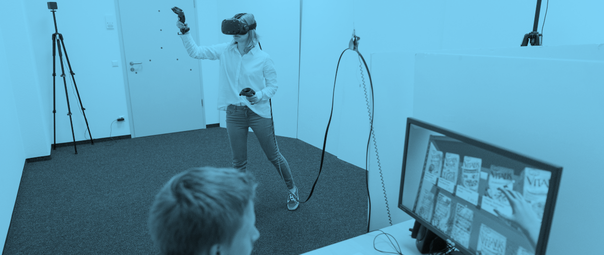 Student mit VR Brille steht im Raum und ein anderer Student sitz vor einem Computer